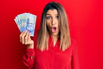 Sticker - Beautiful brunette woman holding south african 100 rand banknotes scared and amazed with open mouth for surprise, disbelief face