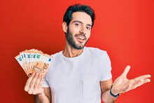 Young Hispanic Man Holding Euro Banknotes Celebrating Achievement With Happy Smile And Winner Expression With Raised Hand