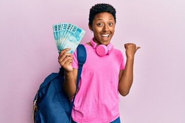 Wall Mural - Young african american girl wearing student backpack and holding brazilian real banknotes pointing thumb up to the side smiling happy with open mouth