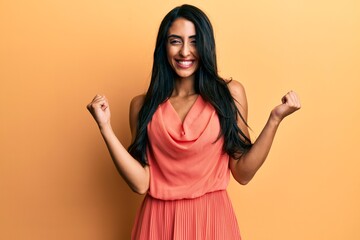 Wall Mural - Beautiful hispanic woman wearing summer dress very happy and excited doing winner gesture with arms raised, smiling and screaming for success. celebration concept.