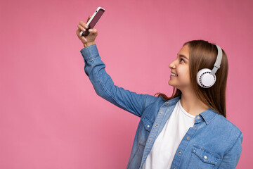 Side-profile Photo of beautiful positive smiling young dark blonde woman wearing blue jean stylish shirt and casual white t-shirt isolated over pink background wearing white bluetooth wireless