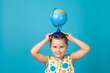 close-up portrait of a smiling charming girl in a white summer dress holding a globe on her head like a hat, draws attention to environmental issues, isolated on a blue background.