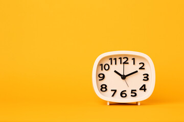 Small white alarm clock, black numbers, set the time placed on a table. Clock on isolated yellow background.