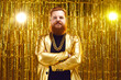 Portrait of bearded guy in funky outfit. Serious unfriendly rich young man in shiny golden disco jacket and gold chain necklace standing arms folded against glittering foil fringe curtain background