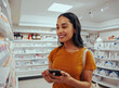 © StratfordProductions - Young woman smiling while using smartphone to read prescription and chose medicine from shelf in pharmacy