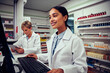 © StratfordProductions - Smiling young woman working in pharmacy using computer with senior colleague using digital tablet in chemist behind counter
