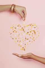 Wall Mural - Shiny heart of falling confetti stars and womans hands on pink background