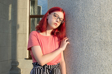 a serious teenage girl with dyed red hair, glasses and freckles. city portrait at sunset.