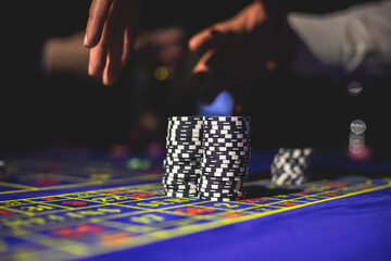 A close-up vibrant image of multicolored casino table with roulette in motion, with casino chips. the hand of croupier, money and a group of gambling rich wealthy people playing in the background