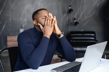 Exhausted African-American Male Employee Covered Face With A Palms, Tired Black Businessman Sits At Workplace With A Laptop, His Eyes Closed, Overwork Concept. Frustrated Man Has Problems With Project