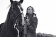 Portrait of a gorgeous brunette woman in an elegant checkered brown jacket posing with a horse on country landscape