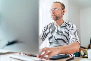A middle-aged man in eyeglasses typing a text using a modern keyboard computer in his living room. Writing or Distance or freelance work on worldwide quarantine time concept