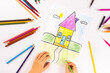 Child girl draws house with multicolored felt-tip pens on a white sheet.