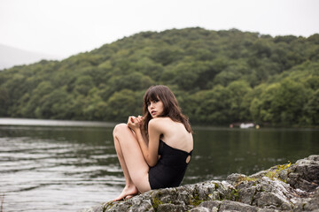 Poster - A beautiful shot of a sexy Caucasian woman wearing a swimsuit and sitting on a rock in front of a lake