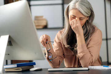 Overworked tired sad senior gray-haired asian woman, freelancer, manager, holds glasses in hand, in stylish clothes, feeling headache, stress after long computer work, massaging eyes at workplace