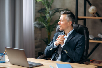 Handsome successful self-confident middle-aged caucasian business leader or manager, sitting in an office in a stylish formal suit, thinking about a project, looking away, taking notes, smiling