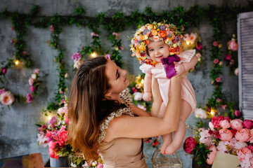 A mother holds her little daughter in her arms with a wreath on her head. Portrait of a beautiful woman and child with a hat made of flowers. Spring decorations.