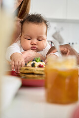 Wall Mural - Cute adorable funny infant mixed race baby child girl playing while multiethnic diverse parents having pancakes on family breakfast sitting at kitchen table at home. Close up view