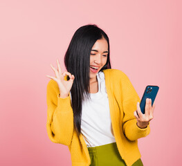 Poster - Happy Asian portrait beautiful cute young woman excited holding mobile phone and gesturing ok sign, studio shot isolated on pink background, Thai female making finger symbol on smartphone