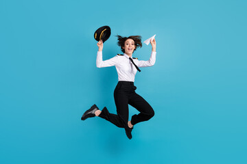 Poster - Full length photo of charming cheerful young woman jump up air hold plane origami hat isolated on blue color background