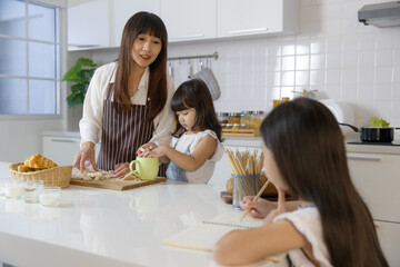 A cute little 7 years old Asian girl sitting and doing homework in kitchen while her mother teaching her younger sister how to make food and cooking behind.