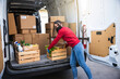 Young female courier unloads van for customer delivery of a basket of fruit and vegetables bought online at supermarket wearing face mask and gloves during global Coronavirus Covid-19 pandemic