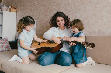 A happy mother teaches her two young daughters to play the acoustic guitar.