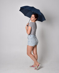 full length portrait of girl wearing shirt and denim shorts, holding an umbrella. isolated on grey studio background.