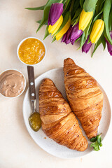 Wall Mural - Breakfast with fresh croissants, chocolate cream, jam and colorful tulips on light stone background. Beautiful romantic composition. Top view.