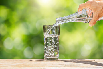 Hand holding drinking water bottle pouring into glass on wooden table on blurred green nature background