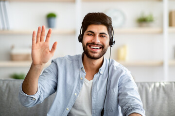 Wall Mural - Greeting concept. Arab man wearing headset and waving hand to camera, gesturing hello and smiling