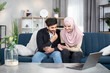© sofiko14 - People, family and technologies concept. Happy young arabian muslim couple using app on mobile phone at home, sitting on blue sofa in living room.