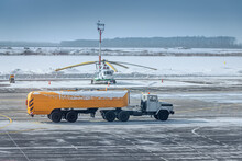 A Fuel Tanker Truck Rides On The Taxiway At The Airport Against The Background Of A Helicopter At Winter Time