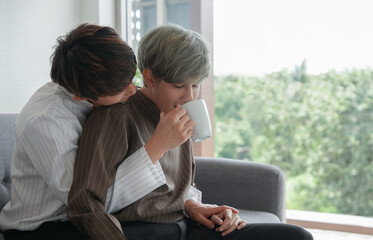 Asian Lgbt men lovers hugging drinking cup of coffee together in the morning. Young man holding cup to feed coffee to boyfriend who sitting on his lap in his arms on sofa near window at home