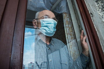 Sad aged man in face mask looking through glazing window surface.