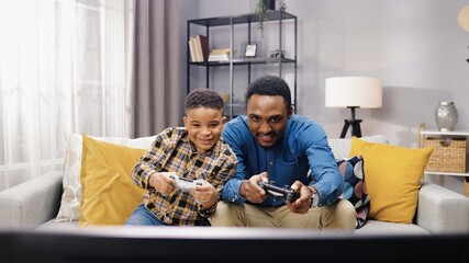 Portrait of African American joyful young male parent having fun with small son sitting on sofa in apartment and playing video games on console in good mood. family time, playtime, gamer concept