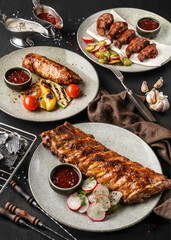 Wall Mural - Grilled pork steak rib and beaf steak meat with vegetables and sauce on plates over dark wooden background. Concept of bbq food, close up