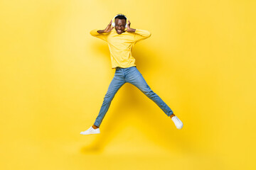 Wall Mural - Happy young African man wearing headphones listening to music and jumping in yellow isolated studio background