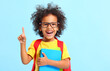 © JenkoAtaman - Happy black kid pointing up while standing in blue studio with school copybooks