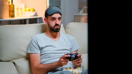 Wall Mural - Close up of man with eye sleep mask playing videogames with joystick for gaming competition. Caucasian male sitting in front on television on sofa late at night in kitchen