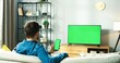 Rear of Caucasian young man in glasses sitting on couch in living room at home watching TV with green screen and typing on smartphone with chroma key browsing online. Social network, tech concept