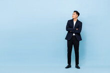 Full Length Portrait Of Young Handsome Southeast Asian Businessman With Arms Crossed Looking Up Sideway To Copy Space On Light Blue Studio Background
