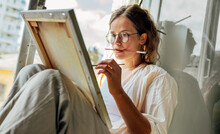 Closeup Portrait Of A Pretty Female Artist Painting On Canvas In Her Art Studio Sitting Next To The Window. A Woman Painter With Eyeglasses Painting With Oil Searching For Imagination In The Workshop.