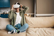 © Drobot Dean - Smiling white woman wearing vr glasses while sitting on sofa at home