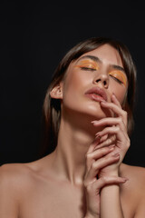 Wall Mural - Close up portrait of artistic young brunette woman with colorful orange makeup posing with eyes closed isolated over black background