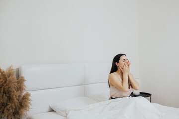 Sleepy girl sitting in bed at early morning and yawns. She covers her mouth with her hands.