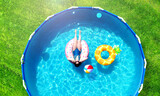 Fototapeta  - Aerial. Girl resting in a metal frame pool with inflatable toys. Summer leisure and fun concept. Frame pool stand on a green grass lawn. Top view from drone.