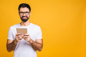 Wall Mural - Happy young bearded man in casual standing and using tablet isolated over yellow background.