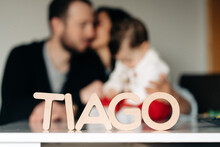 Wooden Tiago Letter Name Placed On Table Near Unrecognizable Young Parent Kissing And Embracing Little Child