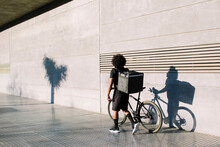 Back View Of Black Delivery Rider With Box And Bike Walking On City Pavement Against Wall With Shadow While Looking Away In Sunlight